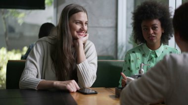 Kahve masasında oturmuş gülen ve gülümseyen üç farklı genç kadın. Konuşurken kız arkadaşlar arasındaki etkileşim. Gerçek hayatta gül ve gülümse.