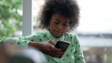 Bir Afro-Amerikalı kadın kahve dükkanında telefon kullanıyor. Akıllı telefon ekranına bakan samimi bir siyahi kadın.