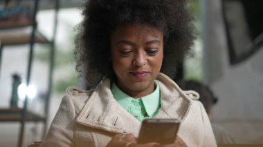 Kahve dükkanında telefonla gülümseyen mutlu bir zenci kadın. Afro-Amerikalı neşeli bir ifadeyle akıllı telefona bakıyor.