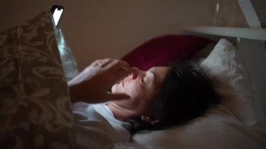 Gece geç saatlerde yatakta uzanmış akıllı telefon cihazına bakan bir kadın.