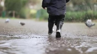 Su birikintisinin içinde koşan küçük çocuk parkta yağmur botları giyiyor.