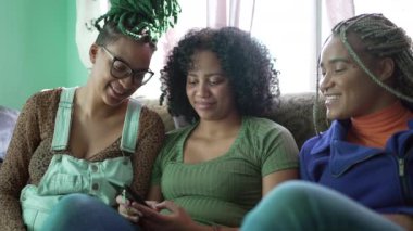 Üç Latin kökenli genç siyahi kadın oturma odasındaki kanepede oturan akıllı telefon cihazına bakıyor. Güney Amerikalı Brezilyalılar modern teknoloji aygıtı kullanıyorlar