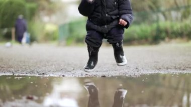 Küçük bebek su birikintisine atlıyor ve çamurlu suda bot yağmuru giyerek koşuyor.