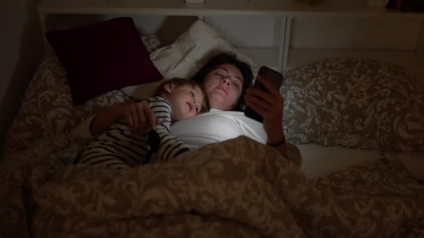 母と子供はベッドで寝そべって夜にスマートフォンを見て — ストック動画