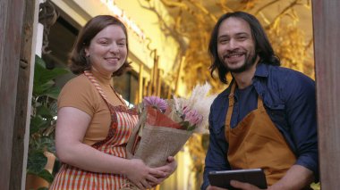 Two employees wearing apron holding tablet smiling at camera. Portrait of workers employees holding flower bouquet arrangement