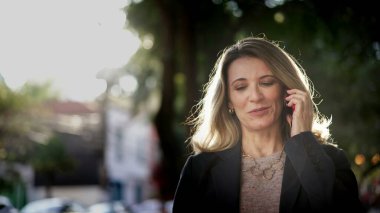 Happy successful middle aged female entrepreneur walking in street talking on phone. Businesswoman speaking on cellphone outdoors in sunlight