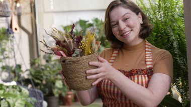 Happy young woman holding bouquet of flower pot showing to camera beautiful arrangement. Portrait girl wearing apron female employee of local small business flower shop