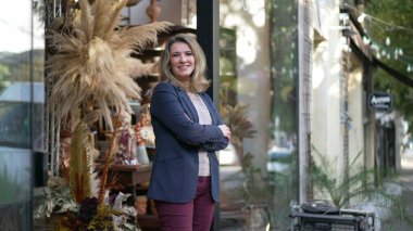 MIddle age entrepreneur smiling standing in the door of her flower shop proud to be the owner of a small business standing by street sidewalk
