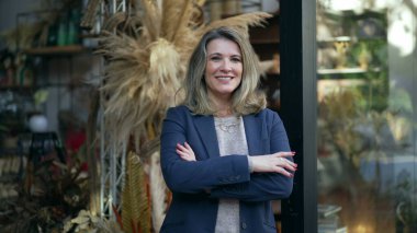 MIddle age entrepreneur smiling standing in the door of her flower shop proud to be the owner of a small business standing by street sidewalk