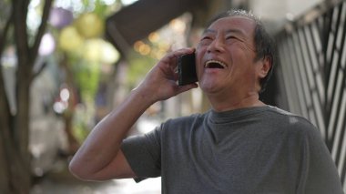 Happy senior man receiving great news on phone standing in city street. Authentic real life laugh and smile of an older Asian person speaking with cellphone laughing