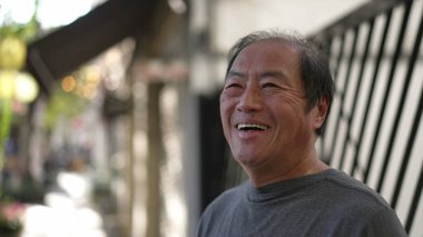 A happy Senior Asian man standing in street sidewalk smiling. Portrait of a middle aged older joyful person in daylight. Authentic real life smile