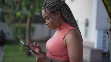 Açık havada duran tablet kullanan genç siyahi bir kadın. Saç stili örülmüş, internetten internette gezinen, elinde ekrana bakan Afro-Amerikalı yetişkin bir kız..