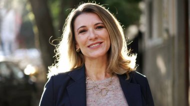 Smiling confident older woman walking outdoors with sun backlight. Proud mature lady walks forward face closeup