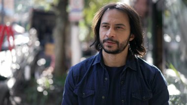 Pensive young casual man walking in street outdoors. Thoughtful person in 30s walks in urban environment. Male with long hair