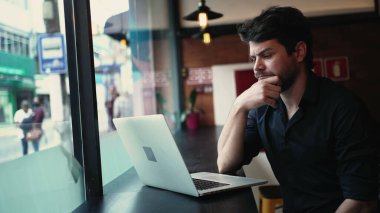 Endişeli genç girişimci, uzak bir iş yerinde, kafenin penceresinde oturan dizüstü bilgisayarın önünde oturuyor.