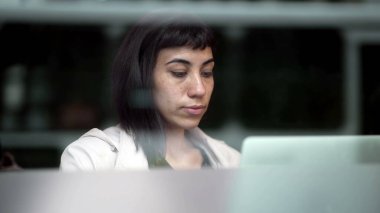 Şehirdeki kaldırım yansıması 2 'ye bakan pencerenin yanında bilgisayarın önünde çalışan genç bir kadın.