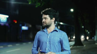 Kaygısız genç adam, yağmurlu bir yağmurda gecenin bir yarısı elinde telefonla şehir kaldırımında yürüyor. Mutlu adam dışarıda yürüyor.
