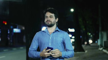 Kaygısız genç adam, yağmurlu bir yağmurda gecenin bir yarısı elinde telefonla şehir kaldırımında yürüyor. Mutlu adam dışarıda yürüyor.