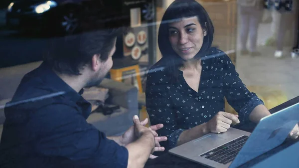 Two colleagues talking about work in front of laptop. Young man pointing at computer screen explaining planning strategy to new female employee seen through window reflection