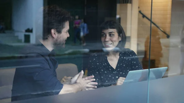 Two young people talking about work in front of laptop and workplace seen through window reflection. Business woman and man discussing in conversation