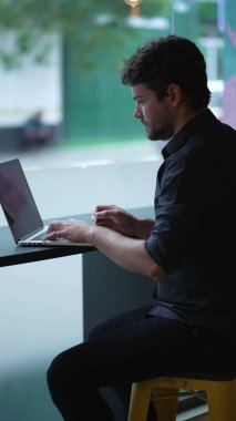 Şehir kaldırımına bakan pencerenin yanında çalışan genç bir adam. Dikey Video 'da dizüstü bilgisayar kullanan düşünceli kişi