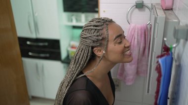 Genç siyahi bir kadın banyo aynasının önünde kendine bakıyor ve dışarı çıkmaya hazırlanıyor. Kutu örgülü Latin Afrikalı bir kız.