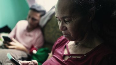 Yaşlı bir İspanyol kadın evde koltukta oturmuş akıllı telefon cihazını kontrol ediyor.