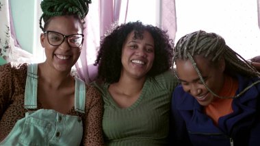 Gülen ve gülümseyen üç kaygısız Brezilyalı genç zenci kadın. Mutlu İspanyol asıllılar gerçek hayatta güler ve gülümserler evde kameraya bakarak.