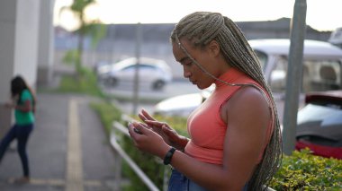 Açık havada tablet kullanan genç bir zenci kadın. Dışarda internette saç stili tarayan İspanyol asıllı bir Brezilyalı.