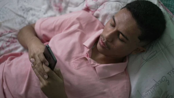 スマートフォンのデバイスにメッセージを入力する1人のヒスパニック系の若者 ベッドに横たわっ電話を使用して男のトップビューの角度 — ストック写真