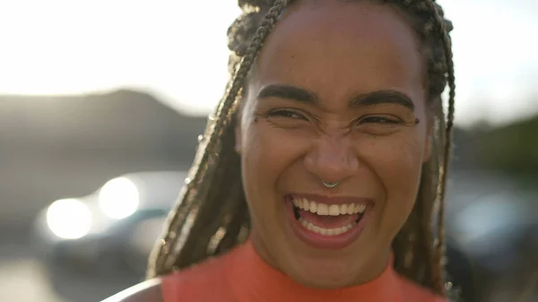 One happy Brazilian young black woman laughing and smiling standing outdoors in sunlight backlight. Happy African American hispanic latin adult girl portrait face closeup