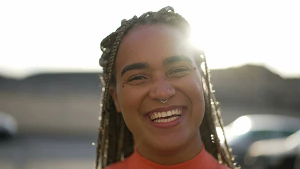 One happy Brazilian young black woman laughing and smiling standing outdoors in sunlight backlight. Happy African American hispanic latin adult girl portrait face closeup