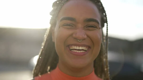 One happy Brazilian young black woman laughing and smiling standing outdoors in sunlight backlight. Happy African American hispanic latin adult girl portrait face closeup