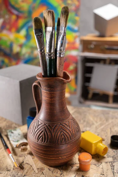 Sanatçı malzemeleri ahşap masada. Çamur sürahisinde boya fırçası ve masa arkası dokusunda resim boyama aracı. Resim için boya fırçası