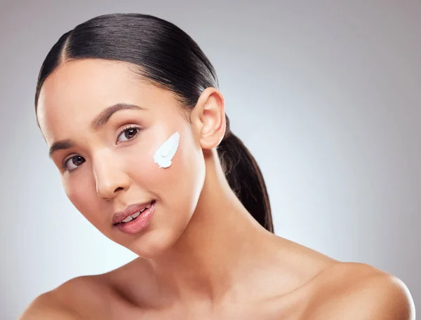 Moisturising helps your skin stay young. Studio portrait of a beautiful young woman applying moisturiser to her face against a grey background. — Stockfoto