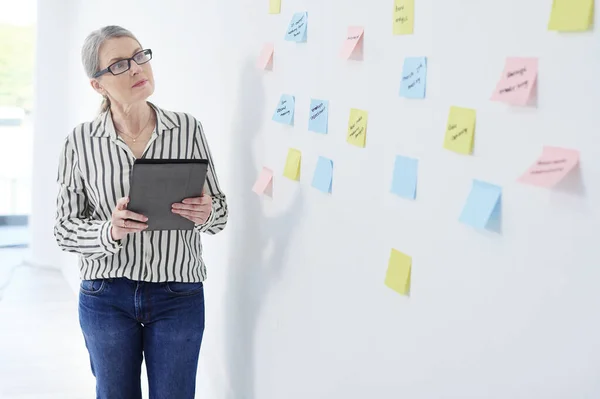 Arbetar mot hennes vision av framgång. Skjuten av en mogen affärskvinna med hjälp av en digital surfplatta medan brainstorming med anteckningar på en vägg i ett kontor. — Stockfoto