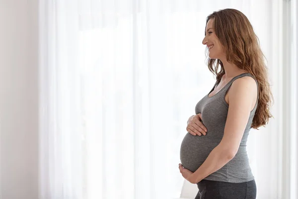 Su bebé está en buenas manos. Foto recortada de una joven embarazada de pie en su casa. — Foto de Stock