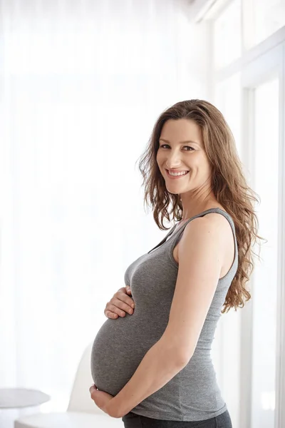 Ενθουσιασμένος με τη μητρότητα. Καλλιεργημένη προσωπογραφία μιας νεαρής εγκύου γυναίκας που στέκεται στο σπίτι της. — Φωτογραφία Αρχείου