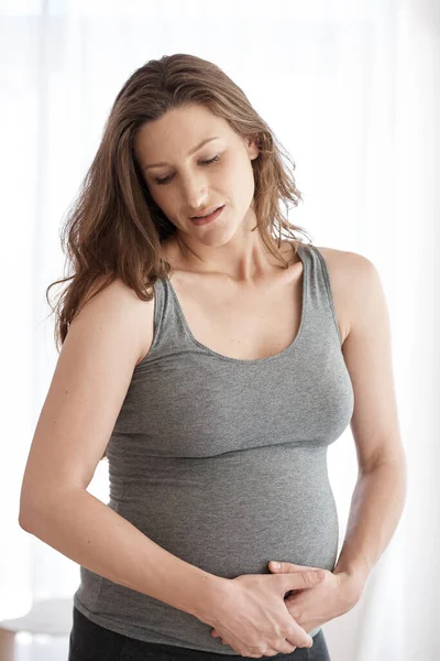 Experiencing some end-of-pregnancy cramps. Cropped shot of a young pregnant woman holding her stomach in pain while standing in her home. — Stock Photo, Image