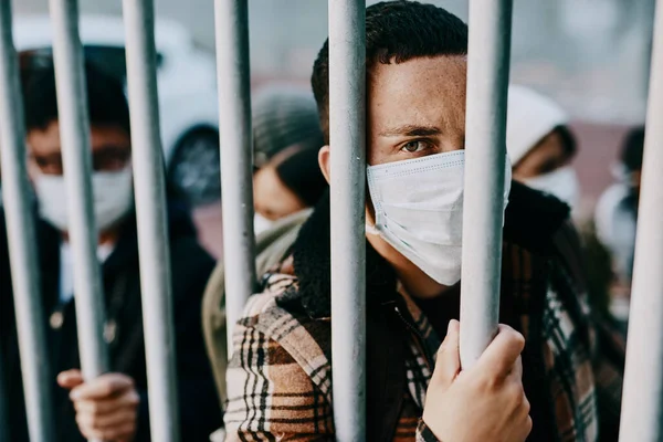 Wer schafft es lebend heraus? Aufnahme eines jungen Mannes mit Maske, der hinter einem Tor in einer fremden Stadt feststeckt. — Stockfoto