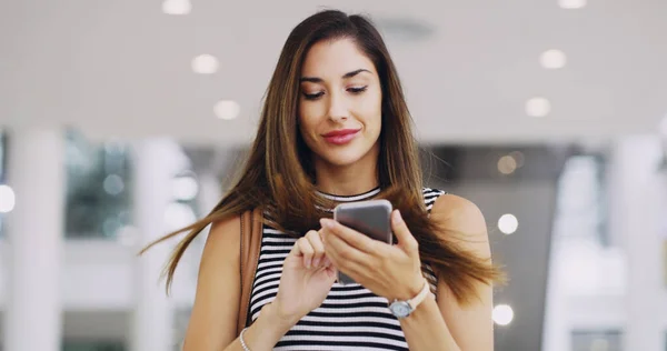 Vale la pena mantenerse conectado. Recorte de una joven mujer de negocios usando un teléfono inteligente mientras camina a través de una oficina moderna. —  Fotos de Stock