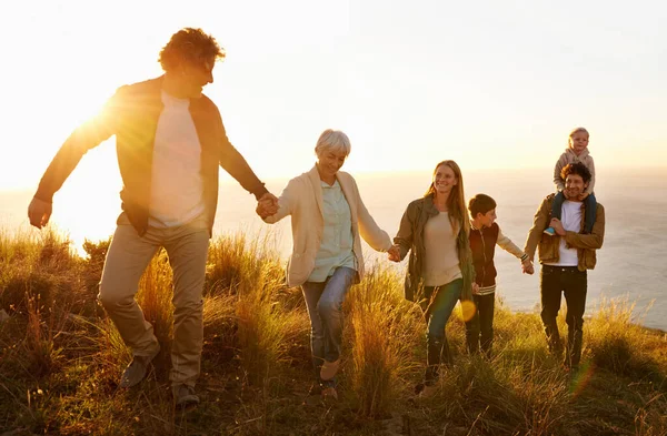 Οικογενειακή ομαδική δουλειά. Μια οικογένεια πολλών γενεών περπατώντας σε ένα καταπράσινο λόφο μαζί το ηλιοβασίλεμα με τον ωκεανό στο παρασκήνιο. — Φωτογραφία Αρχείου