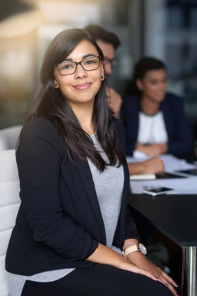 Att lyckas är min högsta prioritet. Porträtt av en leende ung affärskvinna som sitter på ett kontor med kollegor i bakgrunden. — Stockfoto