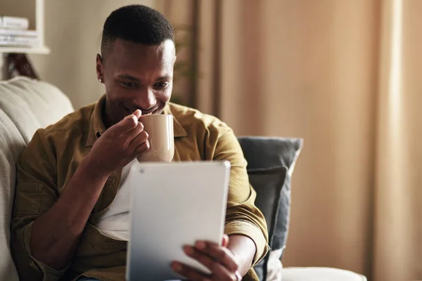 A mídia social é muito interessante hoje. Tiro recortado de um jovem bonito bebendo café enquanto usava um tablet digital em sua sala de estar em casa. — Fotografia de Stock