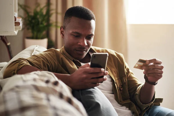 Fazer compras nunca foi tão conveniente. Tiro cortado de um jovem bonito usando um smartphone e um cartão de crédito para fazer compras on-line enquanto sentado em seu sofá em casa. — Fotografia de Stock