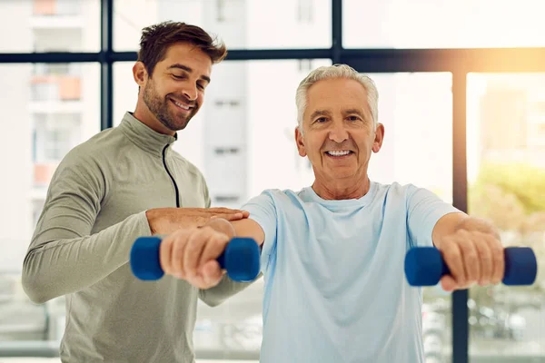 Старі теж можуть бути сильними. Портрет доброзичливого фізіотерапевта, який допомагає старшому пацієнту працювати з вагами . — стокове фото