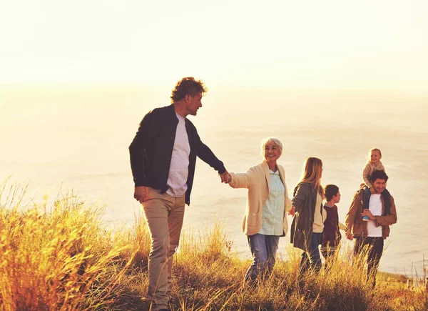 Katakan ya untuk petualangan. Sebuah keluarga multi-generasi berjalan menaiki bukit berumput bersama-sama saat matahari terbenam dengan laut di latar belakang. — Stok Foto