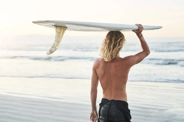 Ma lovagol a hullámokon. Visszapillantás egy fiatal szörfösről, aki a szörfdeszkáját a feje fölött cipeli a parton.. — Stock Fotó