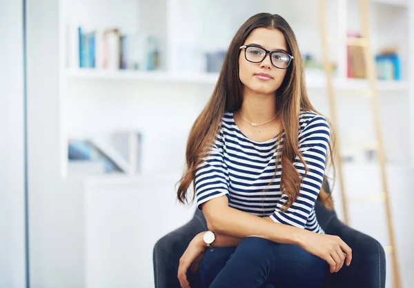 Hon är full av självförtroende. Porträtt av en leende ung kvinna i glasögon som sitter framför bokhyllor hemma. — Stockfoto