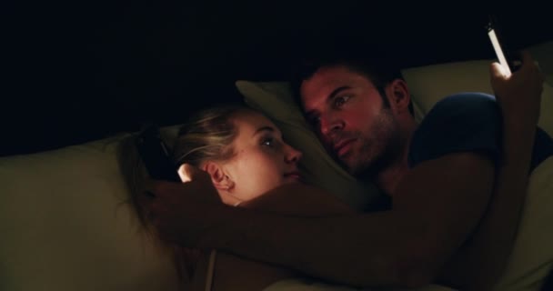Wie verbergt wat voor wie? 4K video beelden van een jong echtpaar controleren van hun telefoons tijdens het knuffelen in bed. — Stockvideo